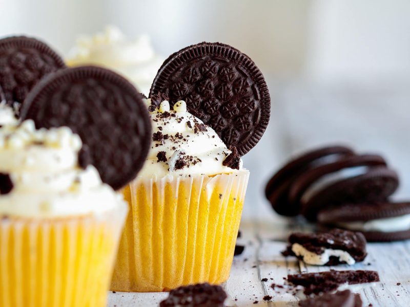 Drei Oreo-Cupcakes in gelben Muffinförmchen auf hellem Holz, daneben liegen ein paar zerbröselte Kekse.