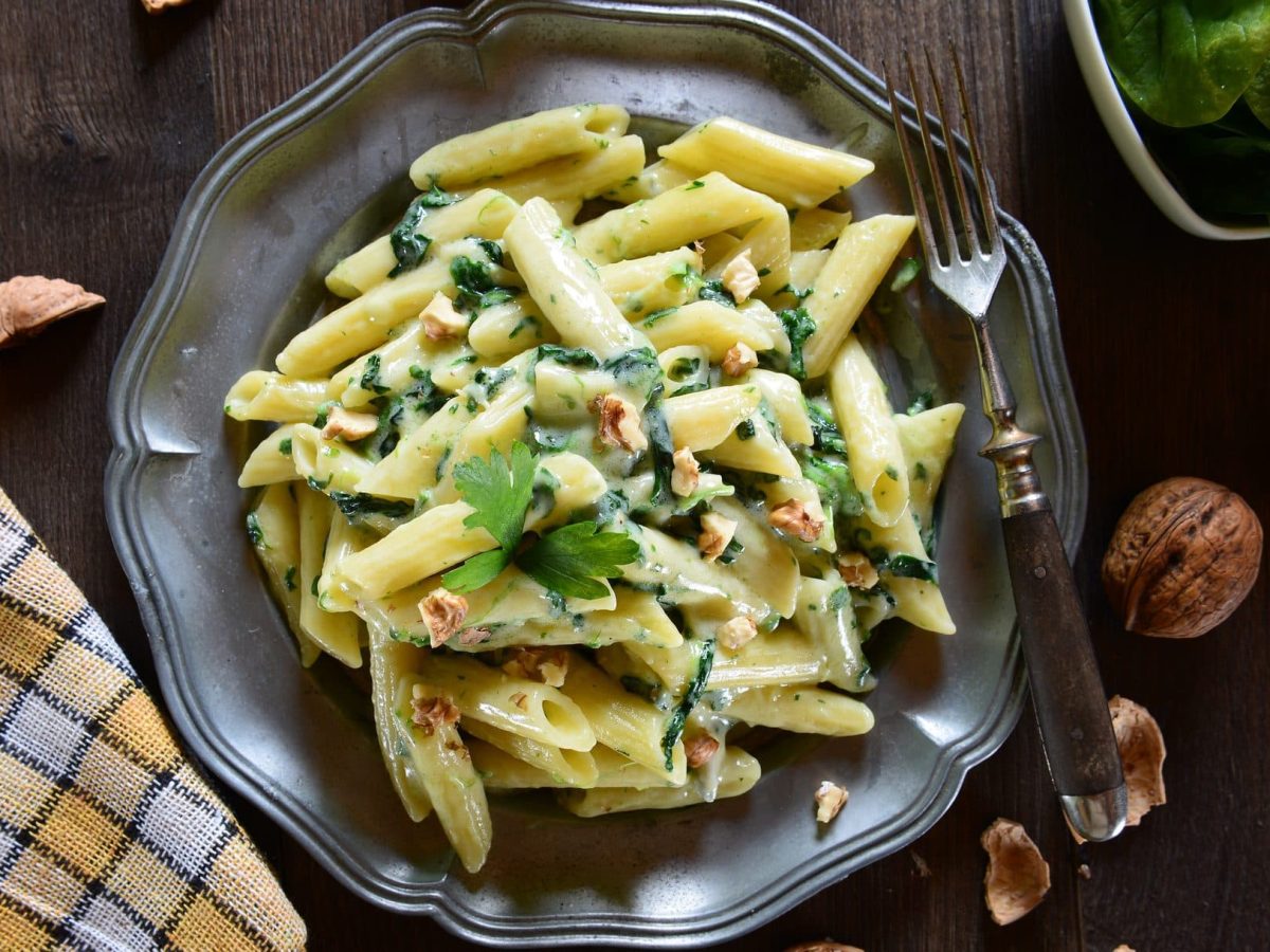 Draufsicht: Auf einem Teller liegen Penne, eine italienische Nudelsorte. Die Penne sind mit Gorgonzola, Spinat und Walnüssen zubereitet.