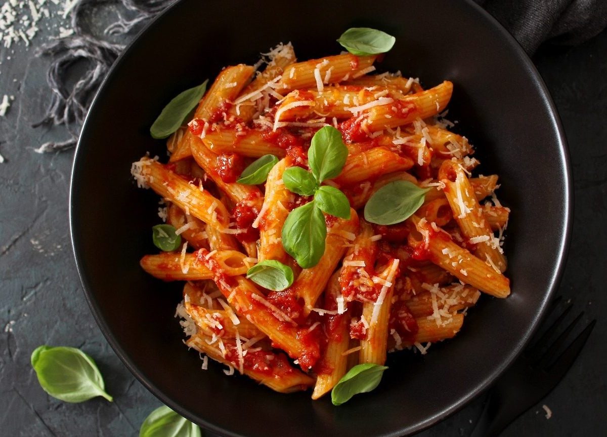 Ein schwarzer Teller mit Penne-Nudeln mit Tomatensauce, Parmesan und Basilikum steht auf schwarzem Hintergrund.