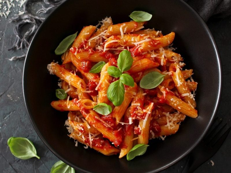 Ein schwarzer Teller mit Penne-Nudeln mit Tomatensauce, Parmesan und Basilikum steht auf schwarzem Hintergrund.