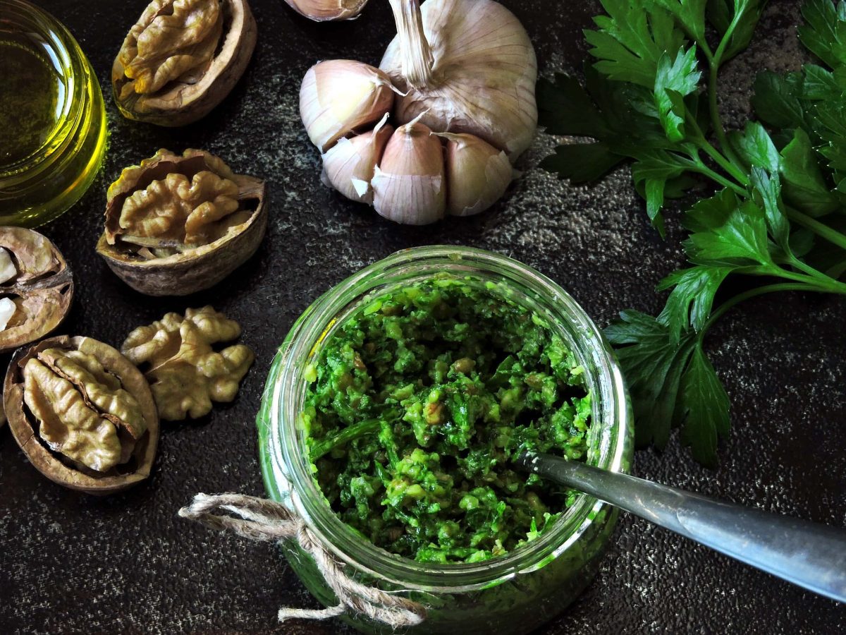 Draufsicht von oben: Auf einem schwarzen Hintergrund sind ein Knoblauch, Walnusshälften, Öl und Petersilienzweige zu sehen. Und in einem Gläschen ist Petersilien-Pesto mit Walnüssen abgefüllt.