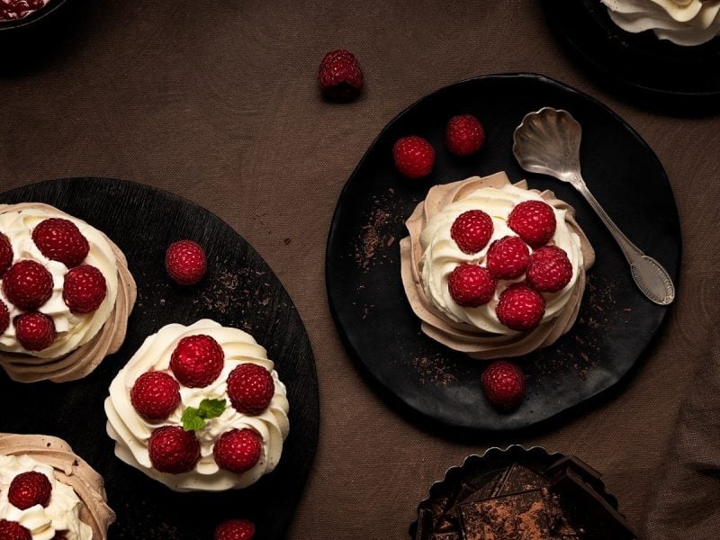 Mehrere schwarze Teller auf denen die Raspberry Meringue inspiriert von Downton Abbey angerichtet sind, neben einem Löffel, weiteren Himbeeren und einer Schale mit Schokolade, vor einem braunen Hintergrund, von oben.