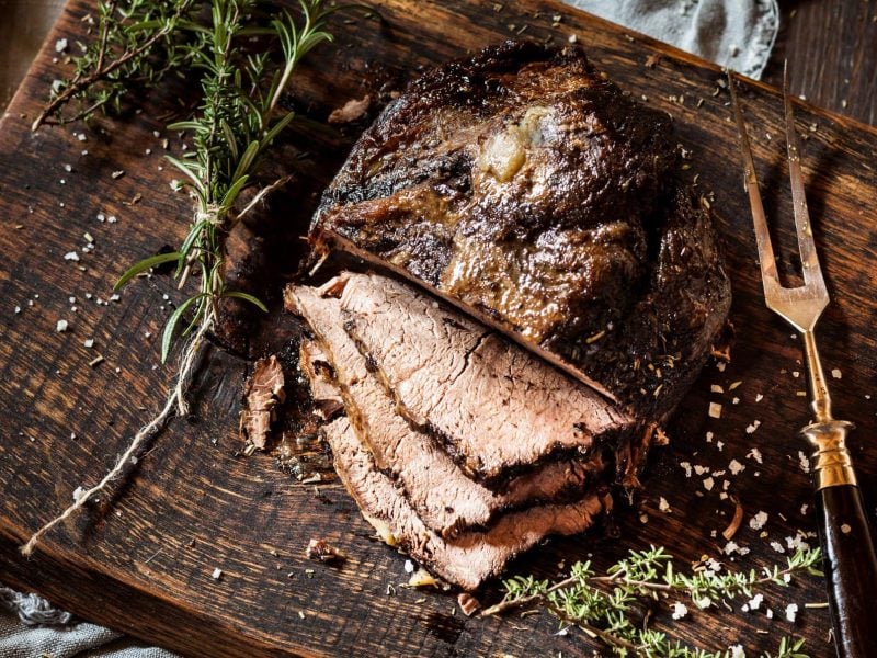 Augeschnittenes Roastbeef mit Pfefferkruste auf einem rustikalen Holzbrett, daneben liegen Kräuter und eine Bratengabel.