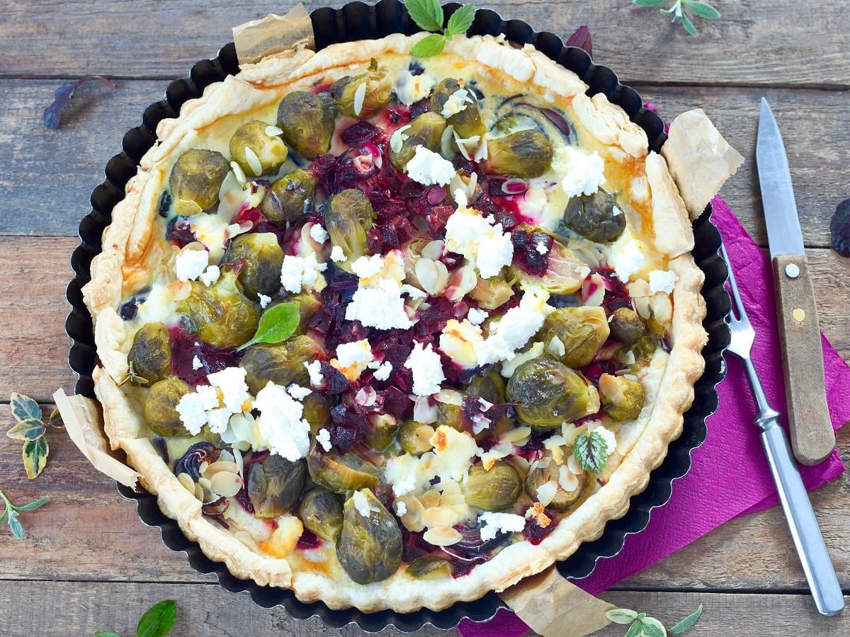 Eine Form mit der frisch gebackenen Rosenkohl-Rote-Bete Quiche mit Ziegenkäse, die mit Mandelblättchen und Kräutern garniert ist und neben der Besteck und eine lila Servierte liegt, vor einem Holzhintergrund, von oben.