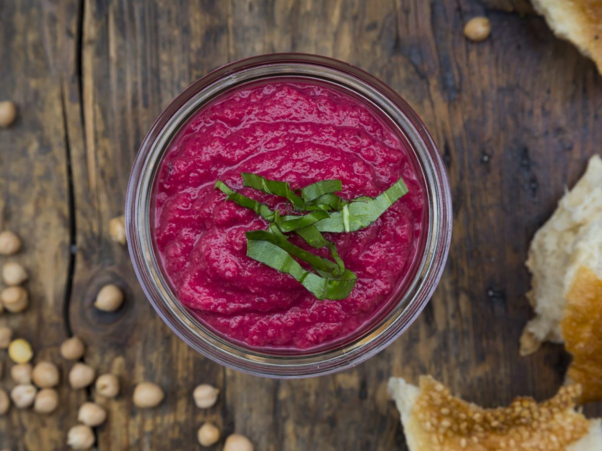 Auf einem rustikalen Tisch steht eine Schale mit Rote-Bete-Hummus. Daneben liegen Kichererbsen und Brot.