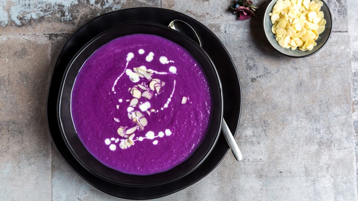 Eine schwarze Schüssel gefüllt mit lila-farbener Rotkohlsuppe die mit einigen Spritzern Sahne und Mandelblättchen versehen ist, neben einem Löffel, einer kleinen Schüssel mit Mandeln, vor einem Steinhintergrund, von oben.