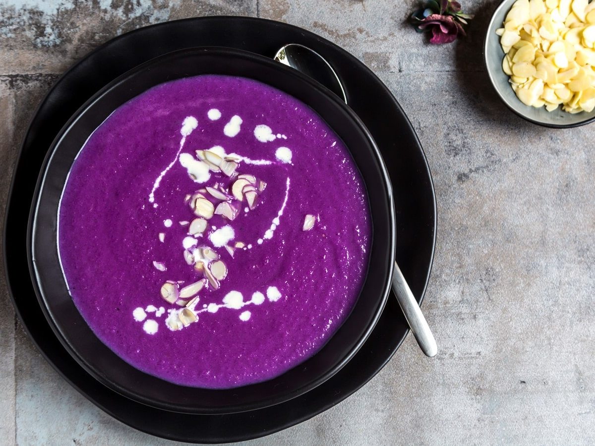 Eine schwarze Schüssel gefüllt mit lila-farbener Rotkohlsuppe die mit einigen Spritzern Sahne und Mandelblättchen versehen ist, neben einem Löffel, einer kleinen Schüssel mit Mandeln, vor einem Steinhintergrund, von oben.