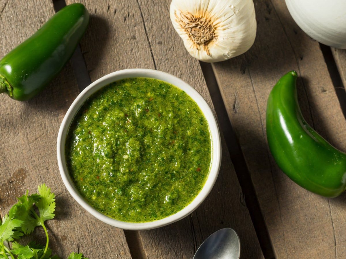 Auf einer Holzplatte liegen zwei grüne Chilischoten, ein Zweig Koriander und Knoblauch. Und eine weiße Schüssel in der eine grüne Paste zu sehen ist. Es handelt sich um die jemenitische Gewürzpaste Zhug.