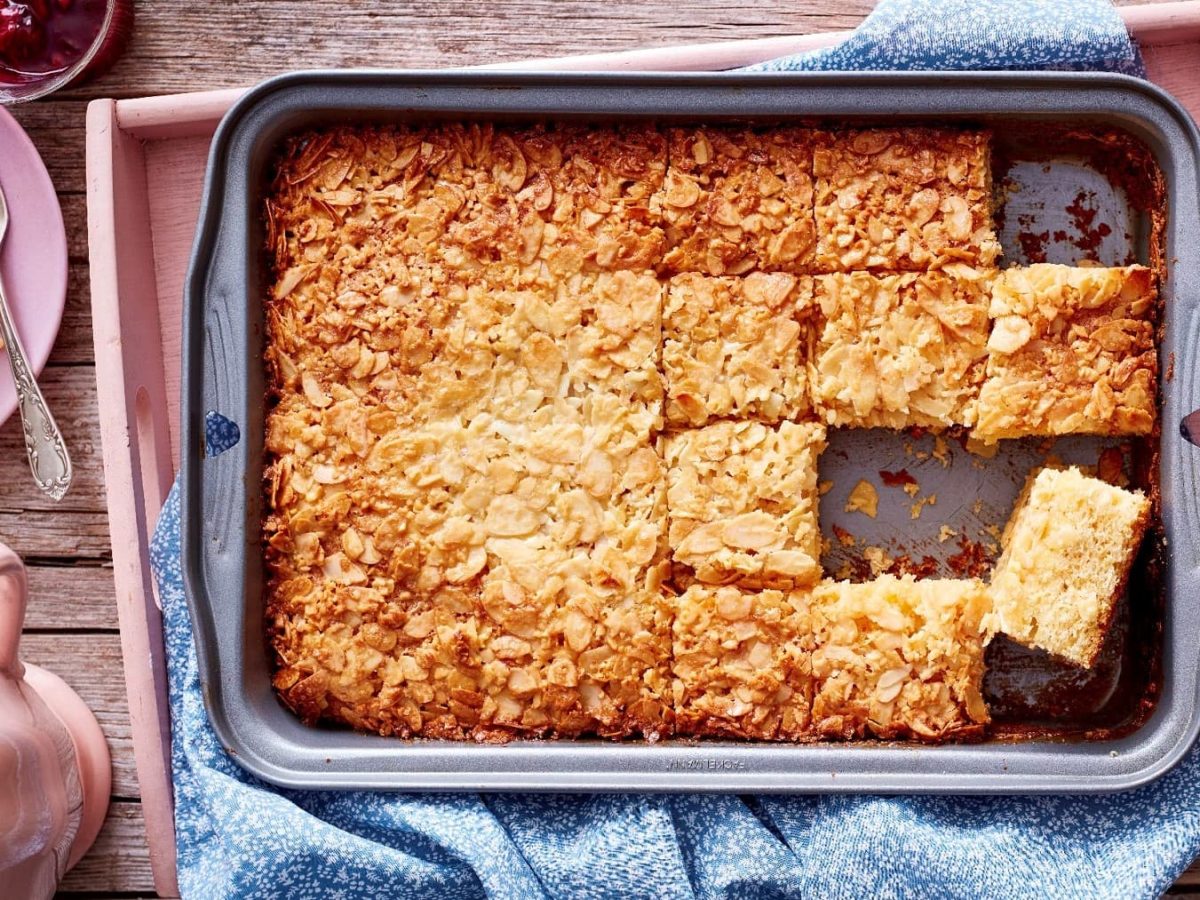 Schneller Butterkuchen in einer tiefen Kuchenform auf einem blau gemusterten Schirrtuch. Die Form steht auf einem rosanen Tablett auf einem hellbraunen Untergrund. Um die Form herum steht eine rosane Teekanne und ein Teller mit einem Stück Kuchen