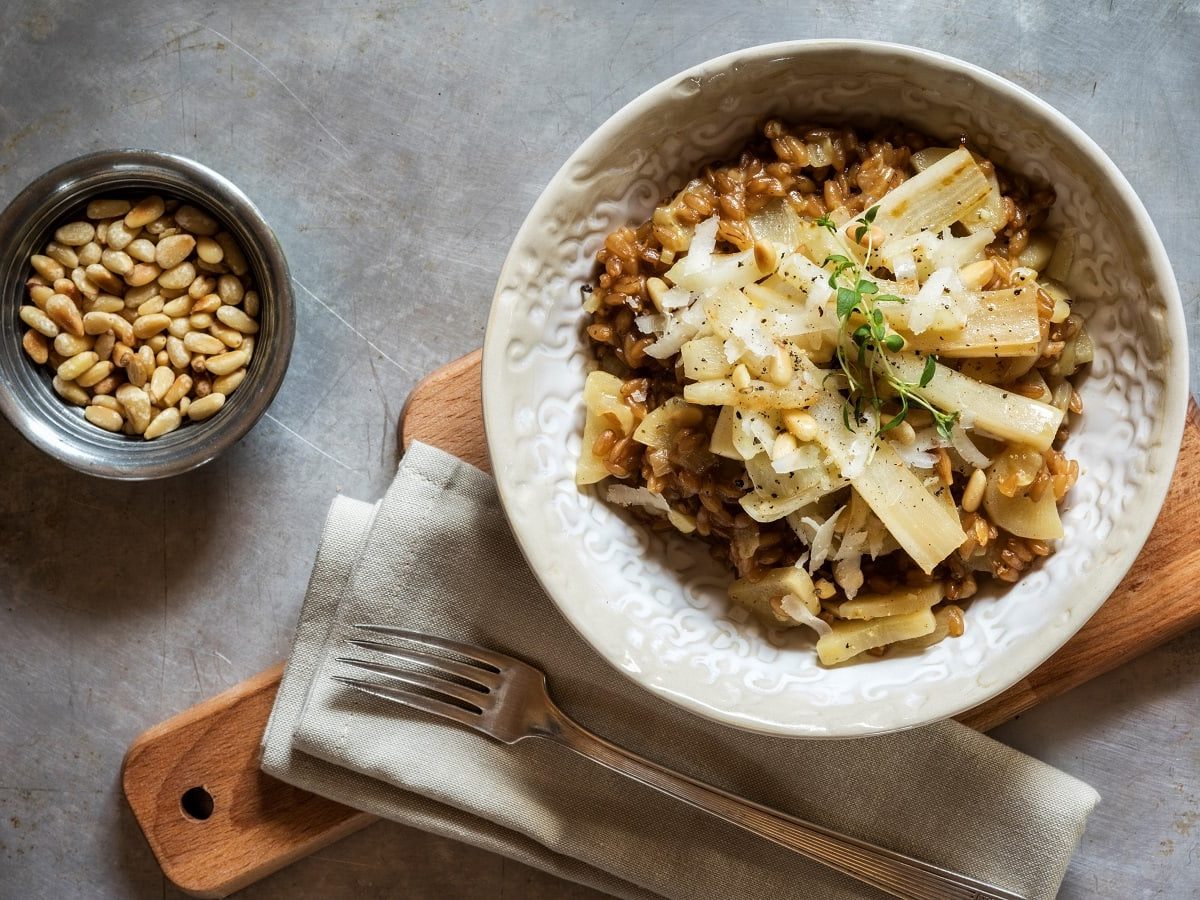 Eine Schale mit einer Portion Schwarzwurzel-Risotto garniert mit Pinienkernen, Parmesan und Kresse auf einem Holzbrett neben einer Stoff-Servierte, einer Gabel und einer Schüssel mit gerösteten Pinienkernen vor einem grauem Hintergrund, von oben.