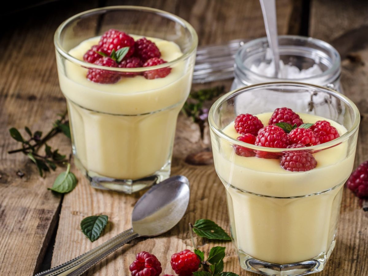 Zwei Gläsern mit Vanillepudding stehen auf einem Tisch. Auf dem Vanillepudding sind Himbeeren und Minzblätter. Auf dem Zisch liegt ein Löffel. Sowie ein flaches Glas mit einem weißem Puder.