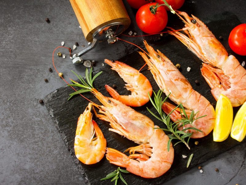 Vier Garnelen liegen auf einer schwarzen Platte. Dazu halbe Zitronenscheiben und zwei Tomaten