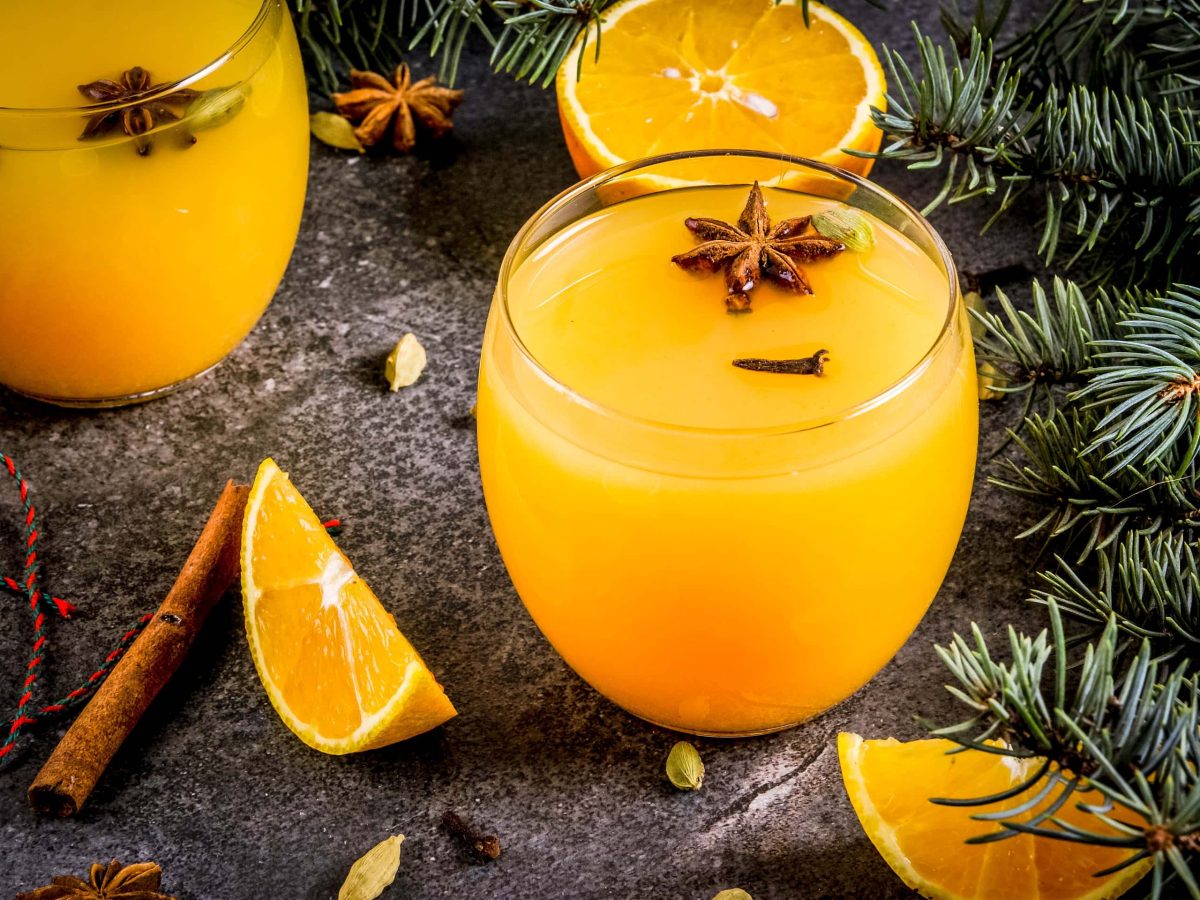 Der Weihnachts-Cocktail mit Orange und Zimt in zwei runden Gläsern angerichtet. Dazu sind im Hintergrund Teile eines Nadelbaums zu sehen.