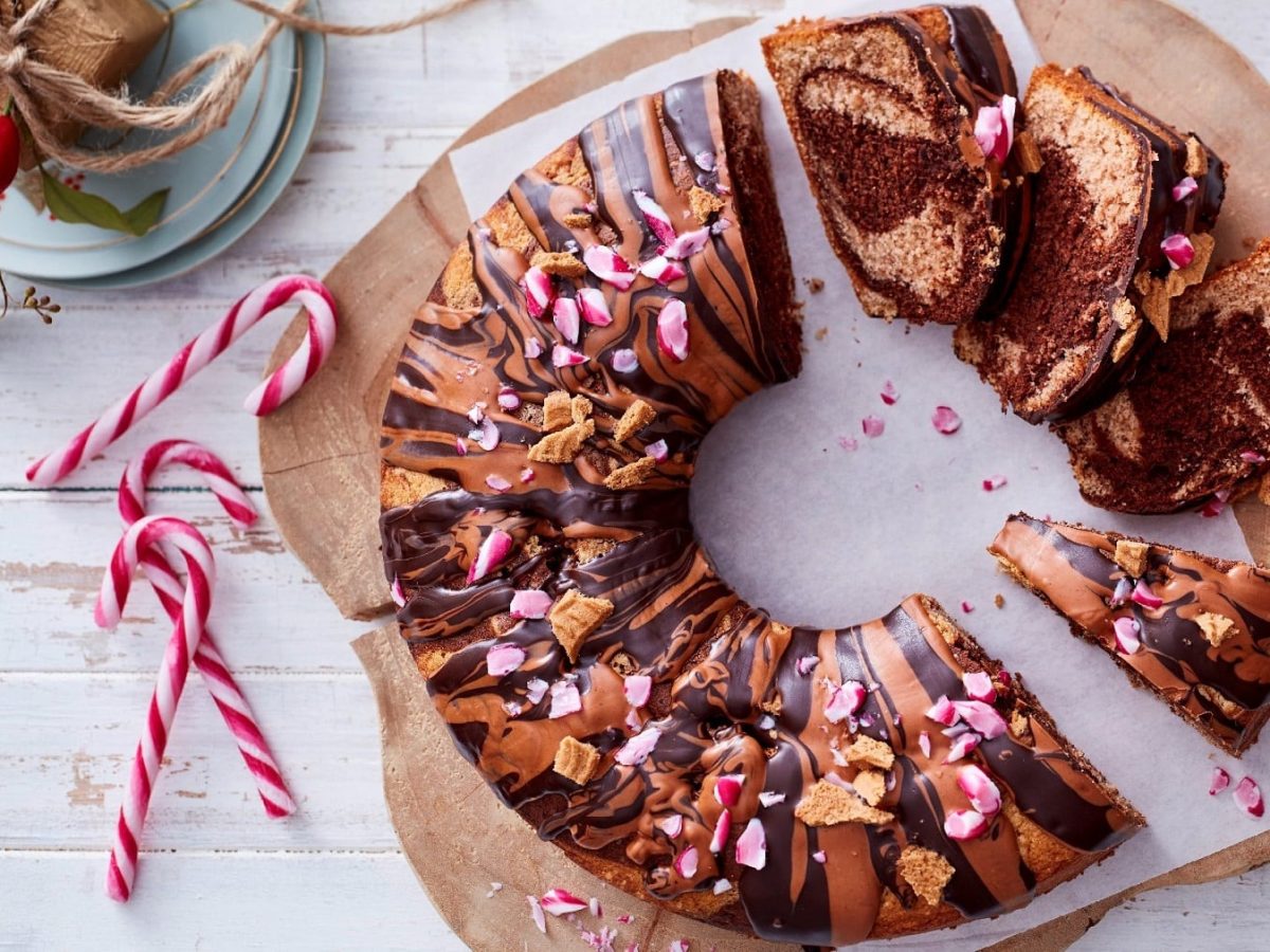 spekulatius-Mamorkuchen angeschnitten auf einem Holzbrett. Der Kuchen ist angerichtet mit Zuckerstangen und Spekulatius-Krümeln. Das Holzbrett steht auf einem weißen Holztisch.