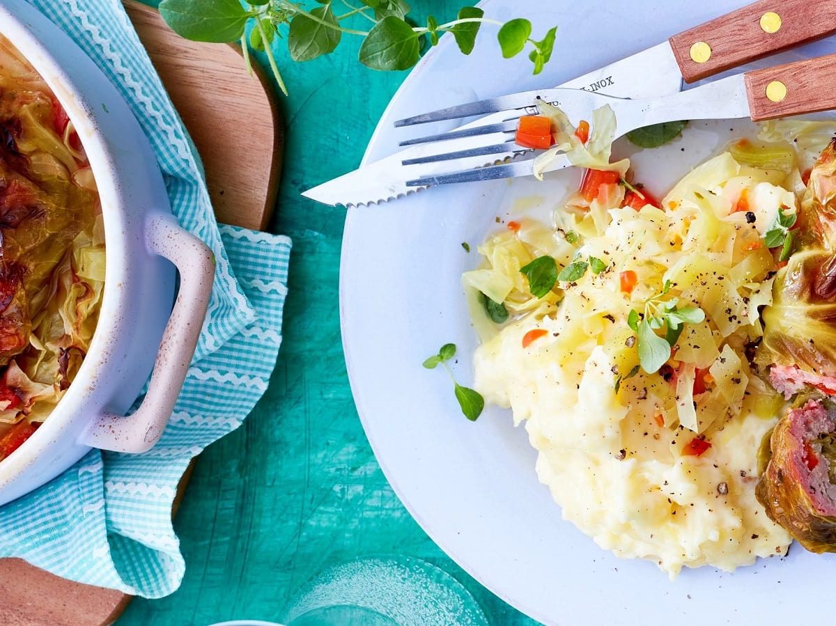 Ein Teller mit einer Portion Spitzkohl-Rouladen mit Gemüse auf Kartoffel-Püree neben Besteck, einem Bräter gefüllt mit weiteren Rouladen, der auf einem Holzbrett steht, und frischem Oregano, vor einem blauen Hintergrund, von oben.