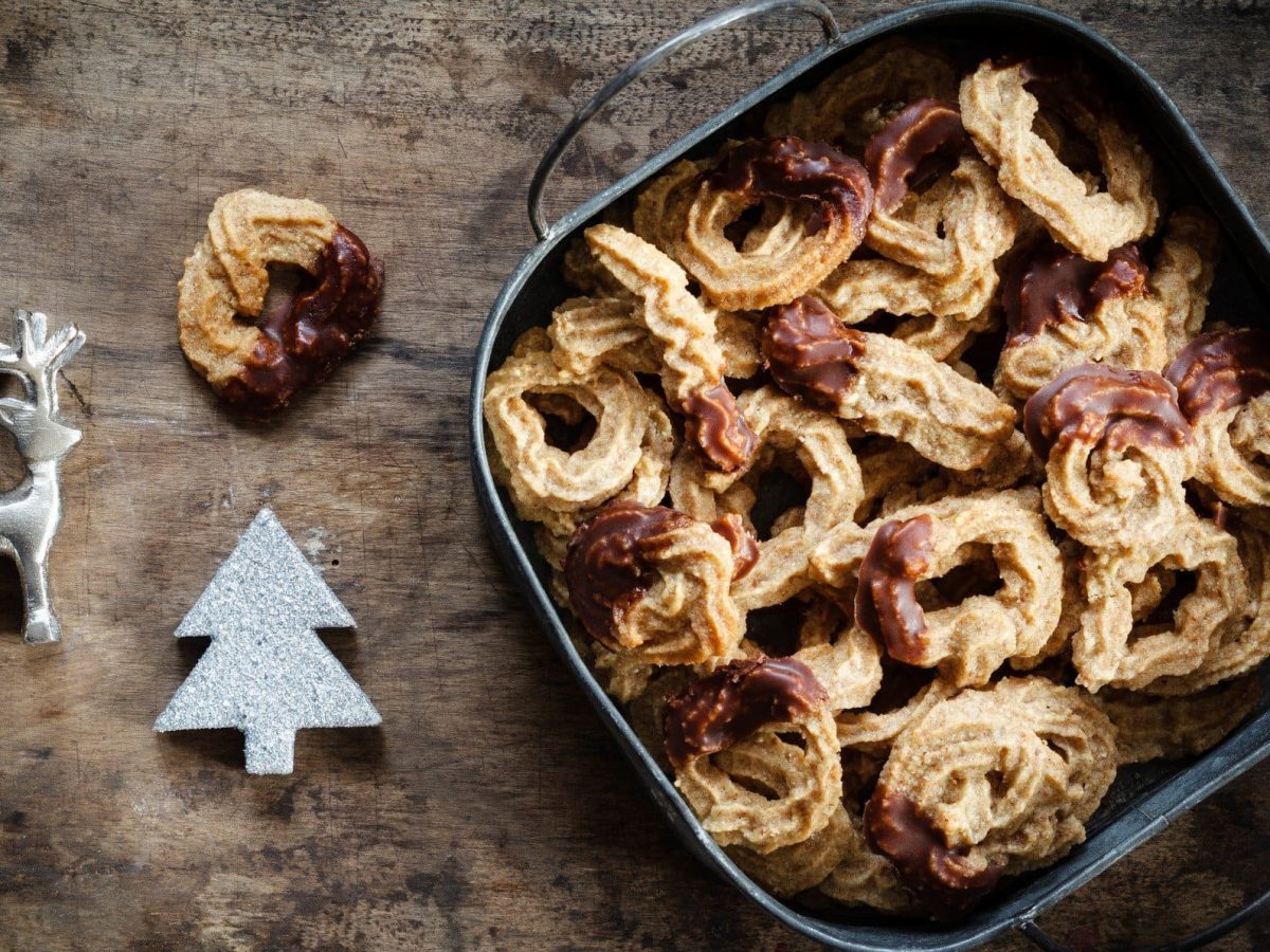 Eine Keksdose mit Spritzgebäck auf dunklem Holz in der Draufsicht, daneben liebt etwas Weihnachtsdeko.