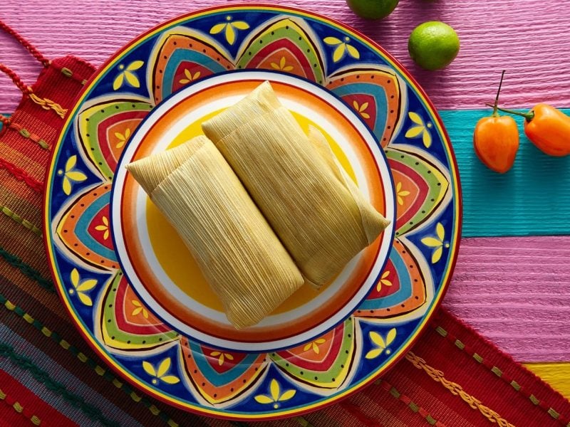 Zwei Tamales mit Schweinefleisch-Füllung auf einem bunt-verzierten Teller vor einem farbigen Teppich und Holzhintergrund neben Paprikaschoten und Limetten, von oben.