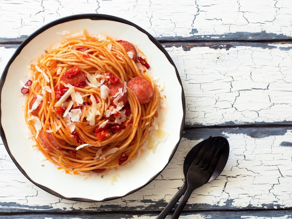 The Big Bang Theory Spaghetti mit Würstchen auf einem weißen Teller mit schwarzem Rand. Der Teller steht auf einem weißen Holztisch. Neben dem Teller liegen ein schwarzer Löffel und eine schwarze Gabel.