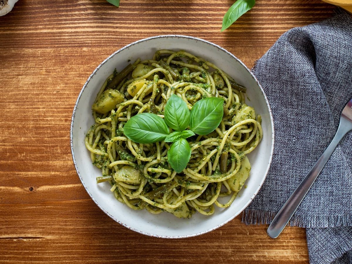 Eine Schüssel mit den Trenette al Pesto gefüllt und garniert mit Basilikumblättern, neben einem grauen Küchentuch auf dem eine Gabel liegt, vor einem Holzhintergrund, von oben.