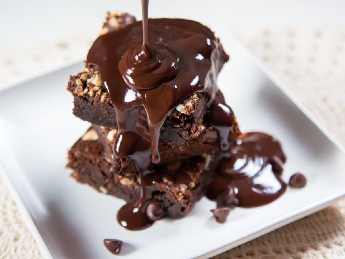 Triple Chocolate Brownie Stücke übereinander gelegt auf weißem Teller und beiger Tischdecke aus der Draufsicht im Moment des Übergießens mit dunkler Ganache.