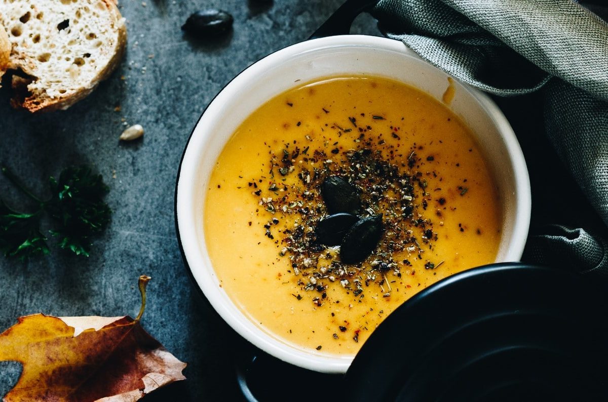 Ein Topf vegane Zucchini-Möhren-Suppe mit Kernen bestreut, daneben liegen zwei Scheiben Baguette und ien paar herbstliche Blätter. Alles in der Draufsicht.