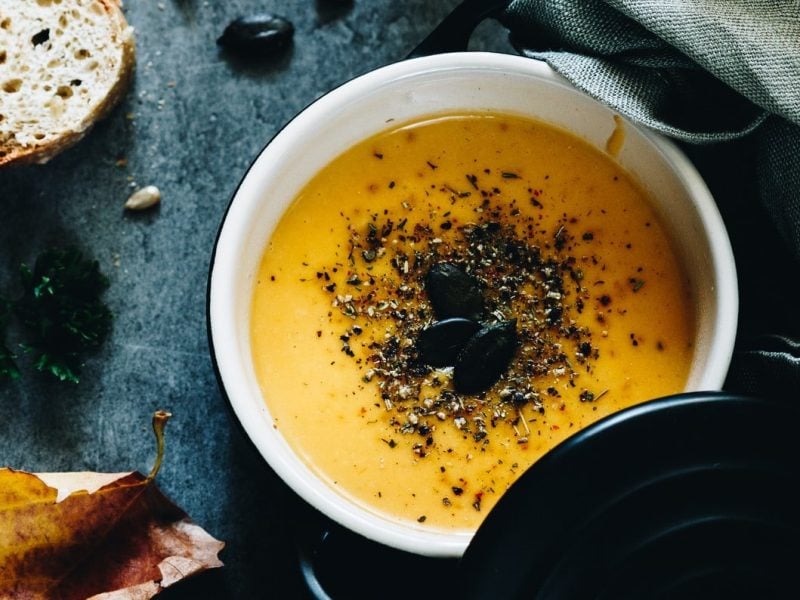 Ein Topf vegane Zucchini-Möhren-Suppe mit Kernen bestreut, daneben liegen zwei Scheiben Baguette und ien paar herbstliche Blätter. Alles in der Draufsicht.