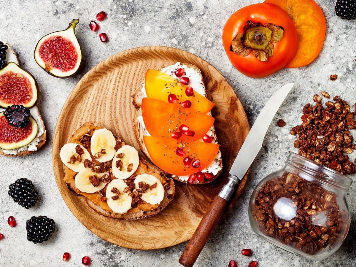 Frühstücksbrot auf einem runden Holzbrett auf einem grauen Untergrund. Ein Brot liegt links neben dem Brett und ist belegt mit Hüttenkäse, Feigen und Brombeeren. Die brote auf dem Brett sind angerichtet mit Erdnussbutter, Banane und Granola und Hüttenkäse, Kaki und Granatapfelkernen. Rechts neben dem Brett liegt ein umgekipptes Glas mit Granola.