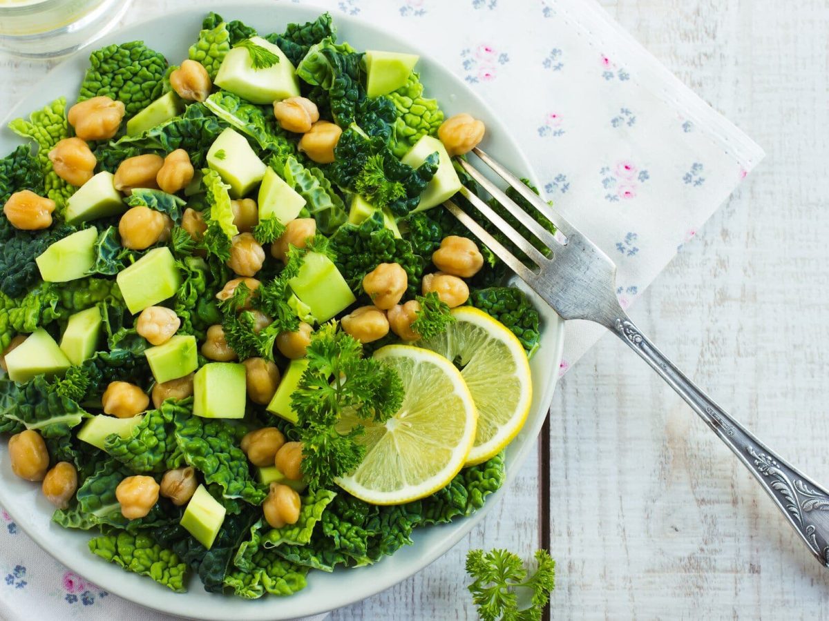 Ein Teller Wirsingsalat mit Kichererbsen und Avocado auf weißem Holz in der Draufsicht mit einer silbernen Gabel.