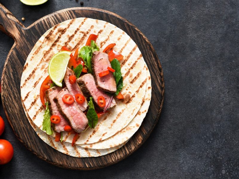 Auf einem runden Holzbrett liegen Wraps mit Roastbeef und Gemüse auf dunkel Untergrund, daneben sieht man Zutaten wie Tomaten und Chilis. In der Draufsicht.