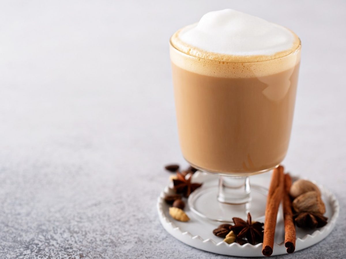 Ein Zimt-Chai-Latte in einem hohen Glas auf einem weißen Untersetzer. Der Untersetzer ist mit Nüssen, Zimtstangen und Anis dekoriert.