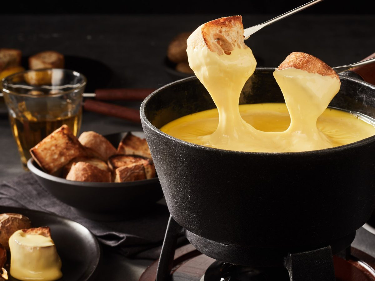 Auf einem Tisch steht ein großer Topf mit Käse-Fondue. Zwei Spieße mit Brot und Käse sind über dem Topf.