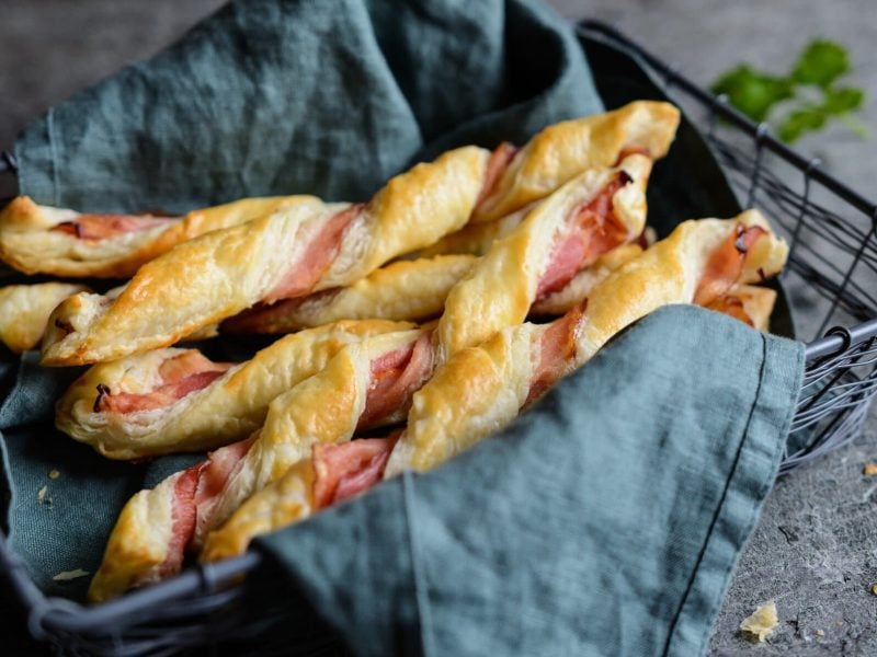 Gedrehte Blätterteigstangen mit Schinken in Brotkorb mit Geschirrtuch.