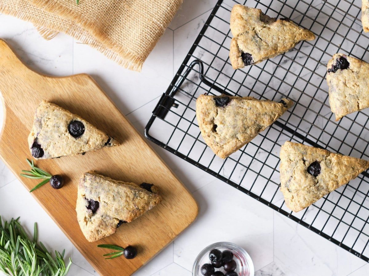 Auf einem Backbleck und einem Holzschneidebrett liegen Blaubeer-Scones. Sie sind dreieckig geschnitten.