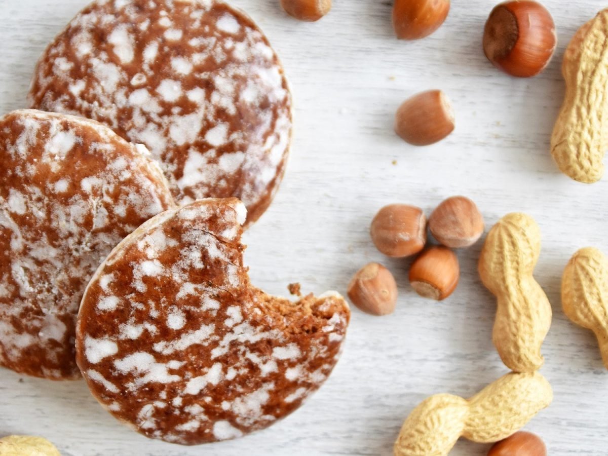 Mehrere Elisenlebkuchen auf einem hellen Untergrund aus Holz. Um die Lebkuchen herum liegen Haselnüsse und Erdnüsse. Einer der Lebkuchen ist bereits angebissen.