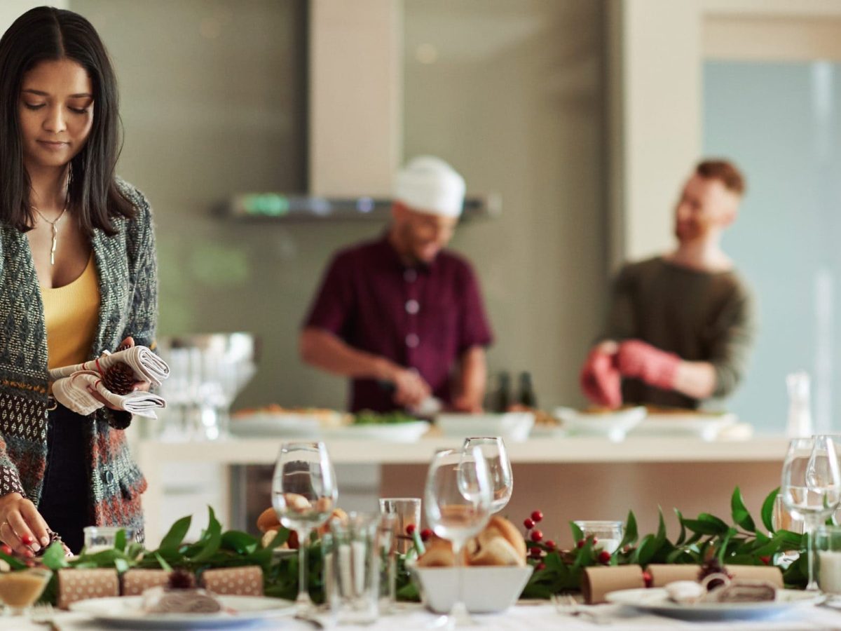 Eine Frau dekoriert den Esstisch, während im Hintergrund zwei Männer in der Küche Essen zubereiten.