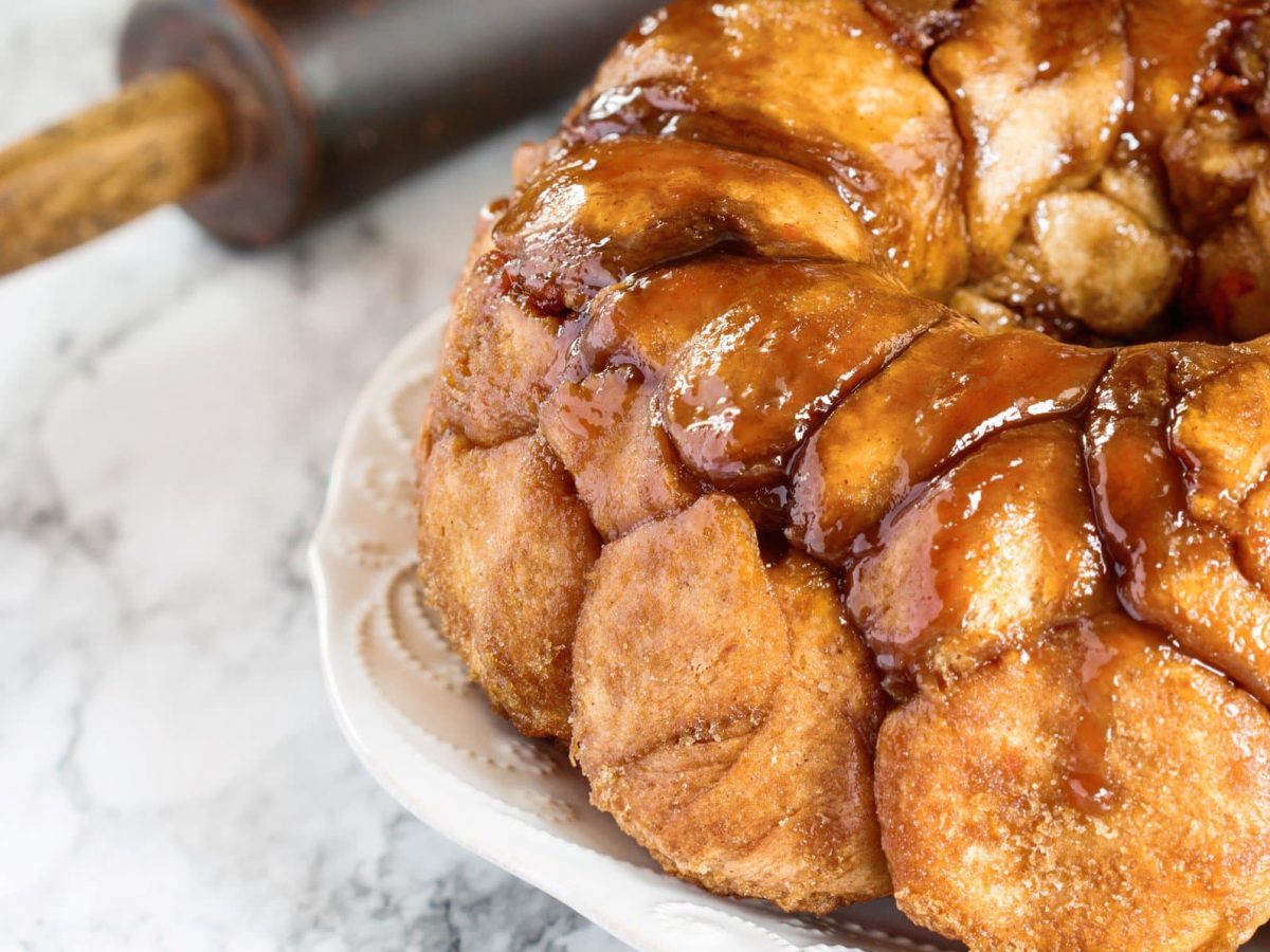 Süßes Monkey Bread mit Karamellsauce übergossen auf weißem Teller auf Marmorplatte. Am linken oberen Bildrand angedeutet schwarzes Nudelholz.