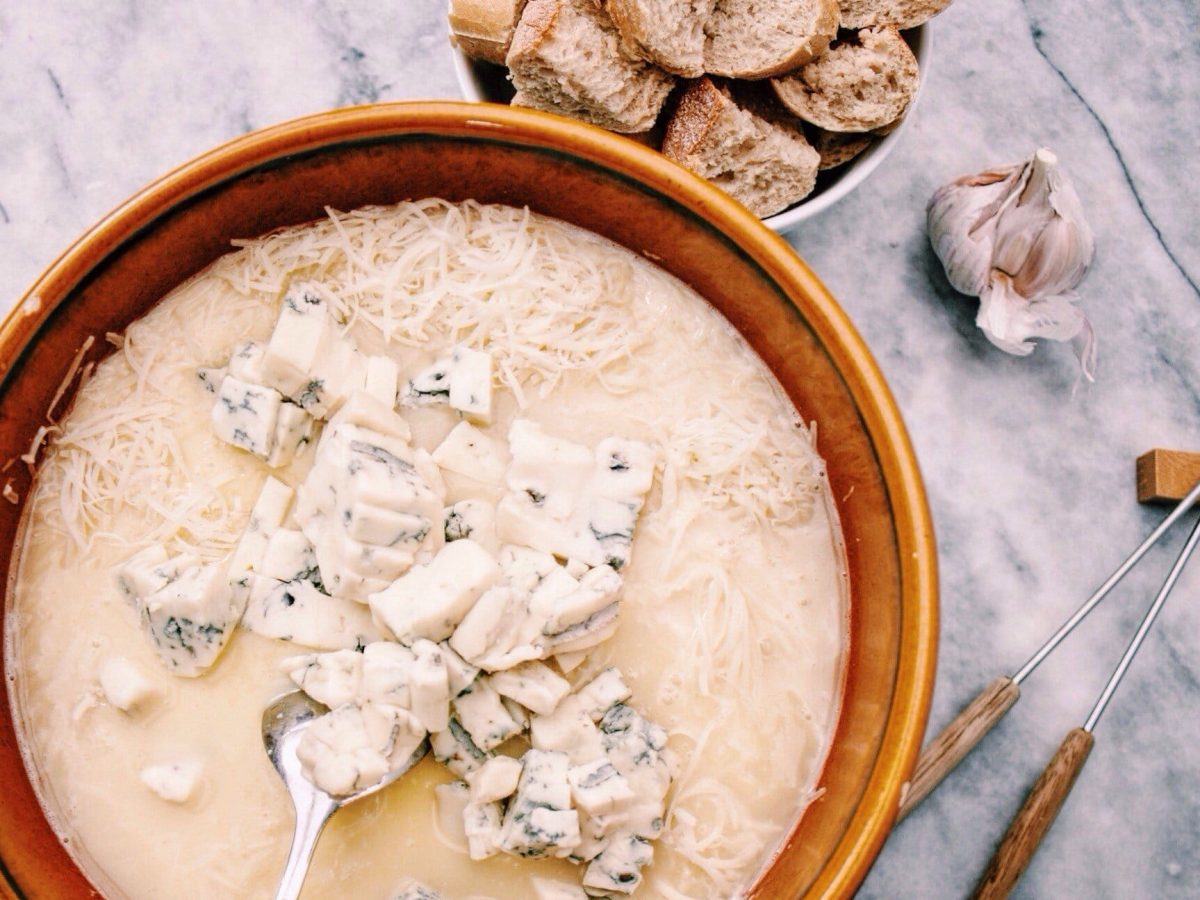 Käsefondue mit Gorgonzola im Tontopf auf Marmoruntergrund. Umgeben ist das Ganz von Brot, Knoblauch und Spießen.