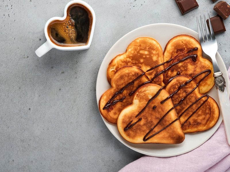 Auf einem Teller liegen herzförmige Buttermilch-Pfannkuchen mit Schokolasdensauce. Daneben steht eine Tasse Kaffee.