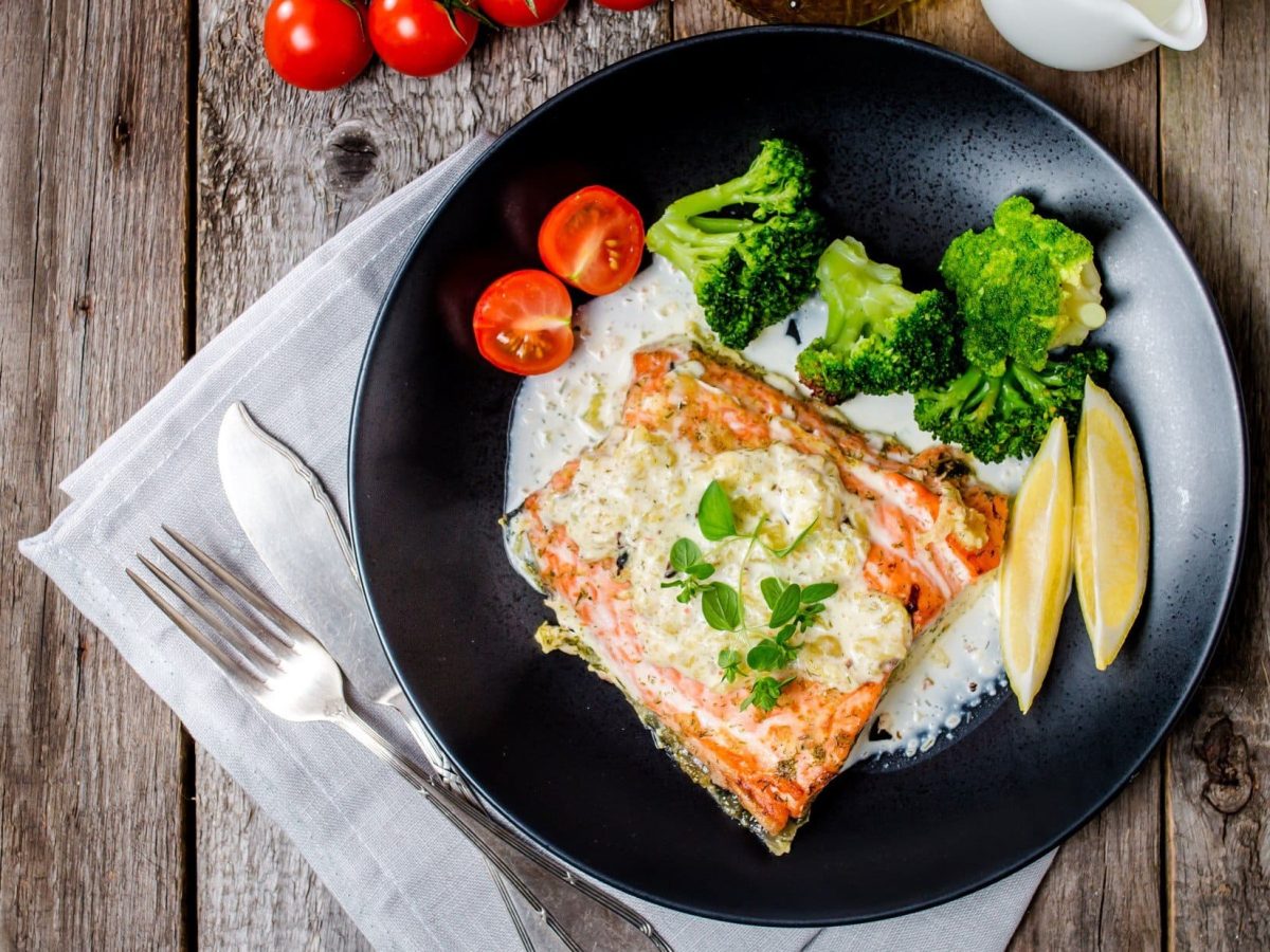 Lachs mit Weißweinsoße und Gemüse auf schwarzem Teller. Dieser steht auf hölzernem Untergurnd.