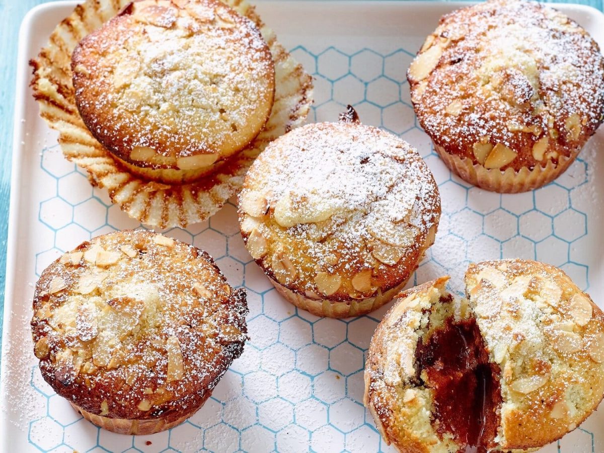 Auf einem weißen Keramikteller liegen 5 Mandel-Muffins mit Espresso-Füllung. Ein Muffin ist angeschnitten, man erkennt im Inneren die Espressofüllung.