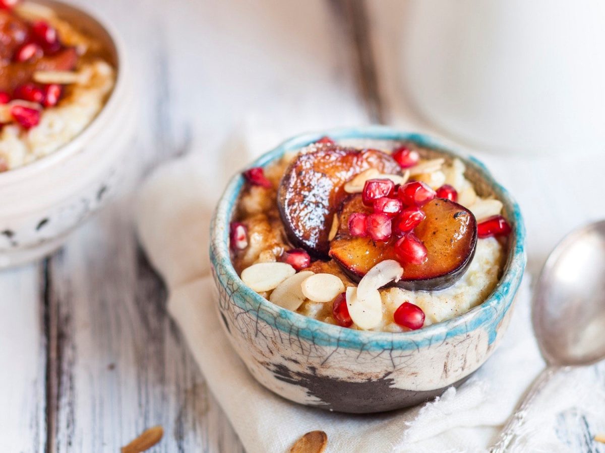 In einer Schüssel ist Pflaumen-Hafer-Bowl. Auf Haferbrei sind Pflaumen, Mandeln und Granatapfel. Daneben liegt ein Löffel.