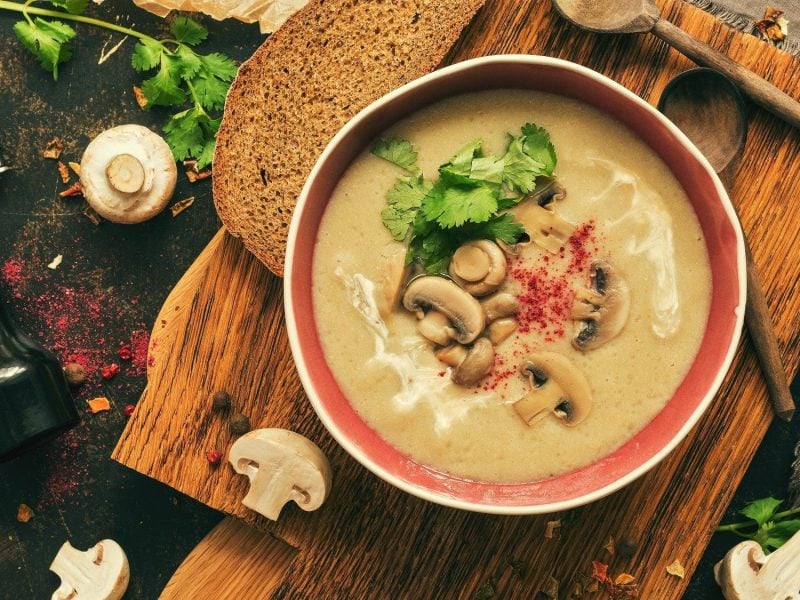 Eine Pilzcremesuppe mit gebratenen Pilzen, Chiliflocken und Petersilie steht auf einem dunklen Holzbrett in einer Schüssel. Um die Schüssel herum liegt Brot, Holzlöffel, Pilze, Petersilie und Gewürze.