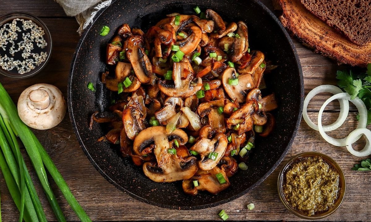 Gebratene Champignons mit Frühlingszwiebeln in Pfanne auf hölzernem Untergrund.