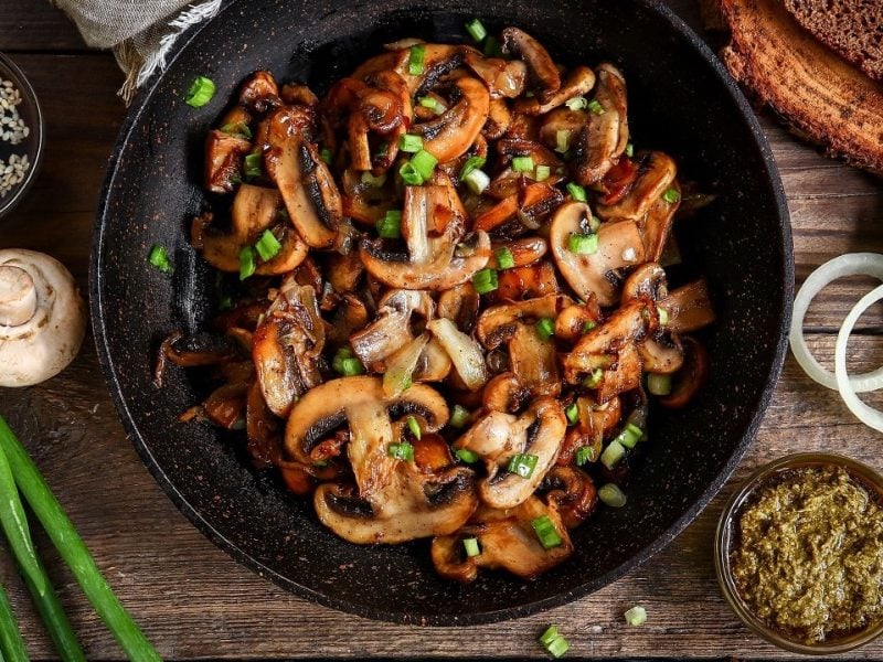 Gebratene Champignons mit Frühlingszwiebeln in Pfanne auf hölzernem Untergrund.