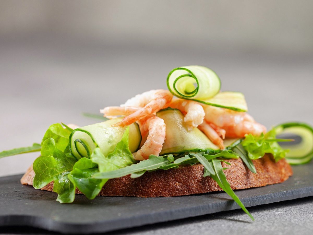 Sandwich mit Garnelen. Das Sandwich steht auf einer schwarzen Schiefernplatte auf einem grauen Untergrund. Das Sandwich ist belegt mit Salat, Gurken und Garnelen.