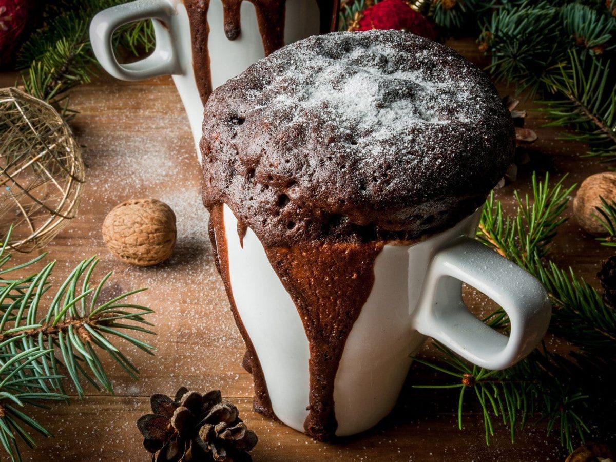 In einer weißen Tasse ist ein schokoladiger Tassenkuchen aufgebacken. Er ist mit Puderzucker bestreut. Die Tasse steht auf einem Holztisch, welcher weihnachtlich dekoriert ist.