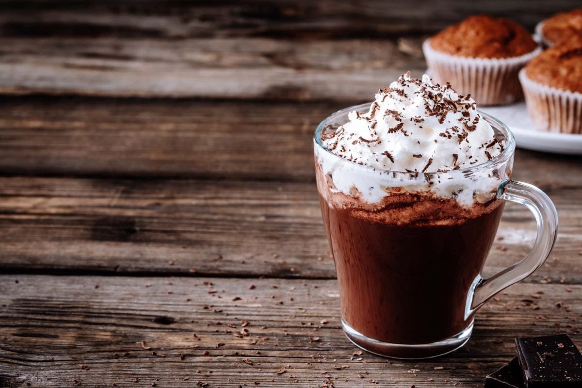 Heißer Kakao mit Rum und Sahne auf hölzernem Untergrund. Im Hintergrund sieht man Muffins.