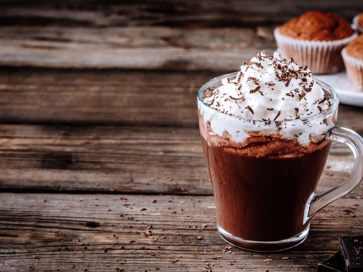 Heißer Kakao mit Rum und Sahne auf hölzernem Untergrund. Im Hintergrund sieht man Muffins.