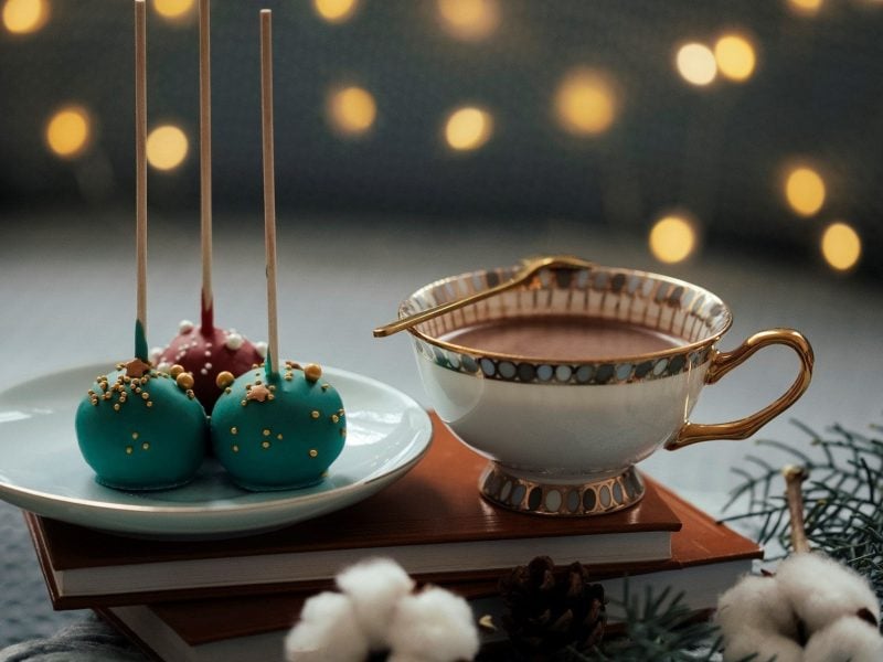 Auf einem Buch steht ein Teller mit weihnachtlichen Cake-Pops und eine Tasse Kakao. Die Cakepops stehen umgedreht auf dem Teller. Die Cake-Pops sind grün und rot gefärbt und mit Zuckerperlen dekoriert.