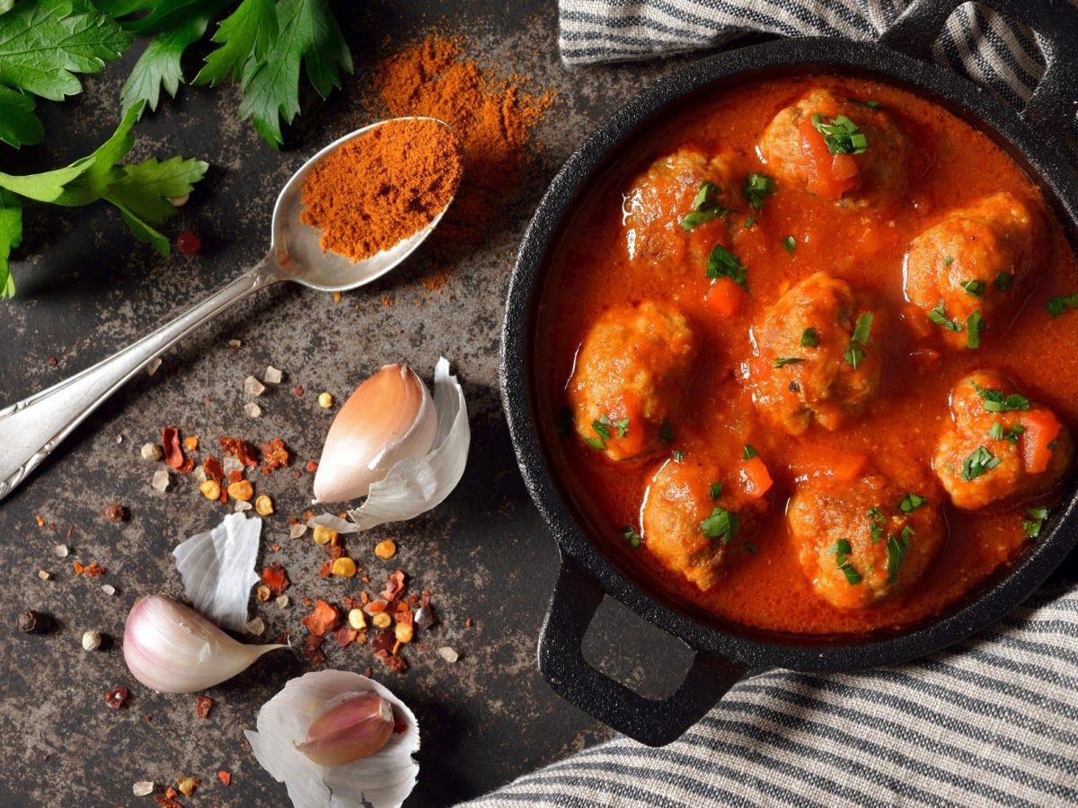 Albóndigas con salsa de tomate in schwarzer Gussform mit Gewürzen und Teelöffel mit Paprikapulver aus der Vogelperspektive. Glatte Petersilie im linken oberen Bildrand. Recht dunkel-hellrau gestreifte Stoffserviette. Arrangiert auf dunklem Holz.